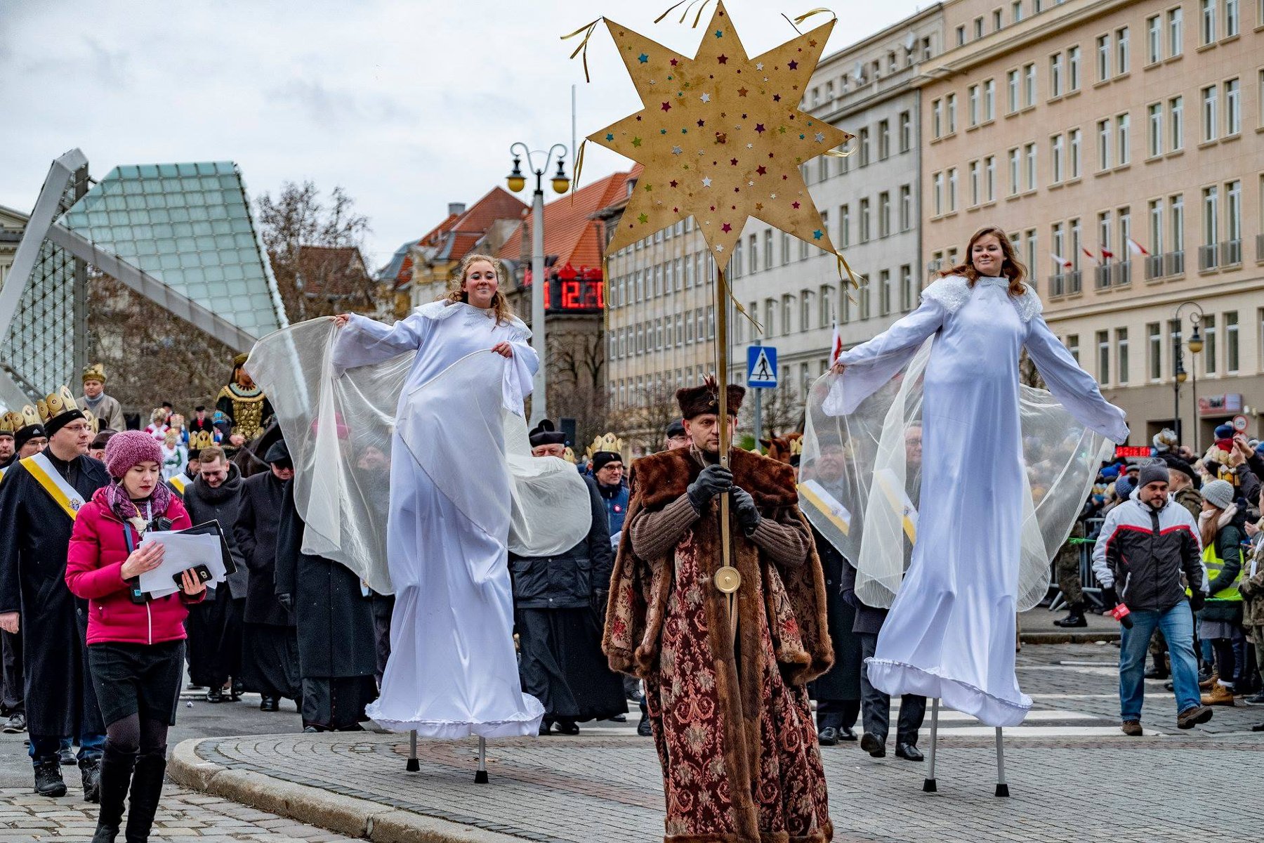 Zdjęcie przedstawia tłum ludzi idących ulicą. W pierwszym rzędzie widać dwie dziewczyny idące na szczudłach w przebraniu aniołów oraz mężczyznę trzymającego w ręku dużą złotą gwiazdę.
