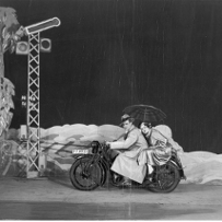Para na motocyklu w scenicznej scenografii skał i drzew. Kobieta trzyma parasol, pada deszcz.