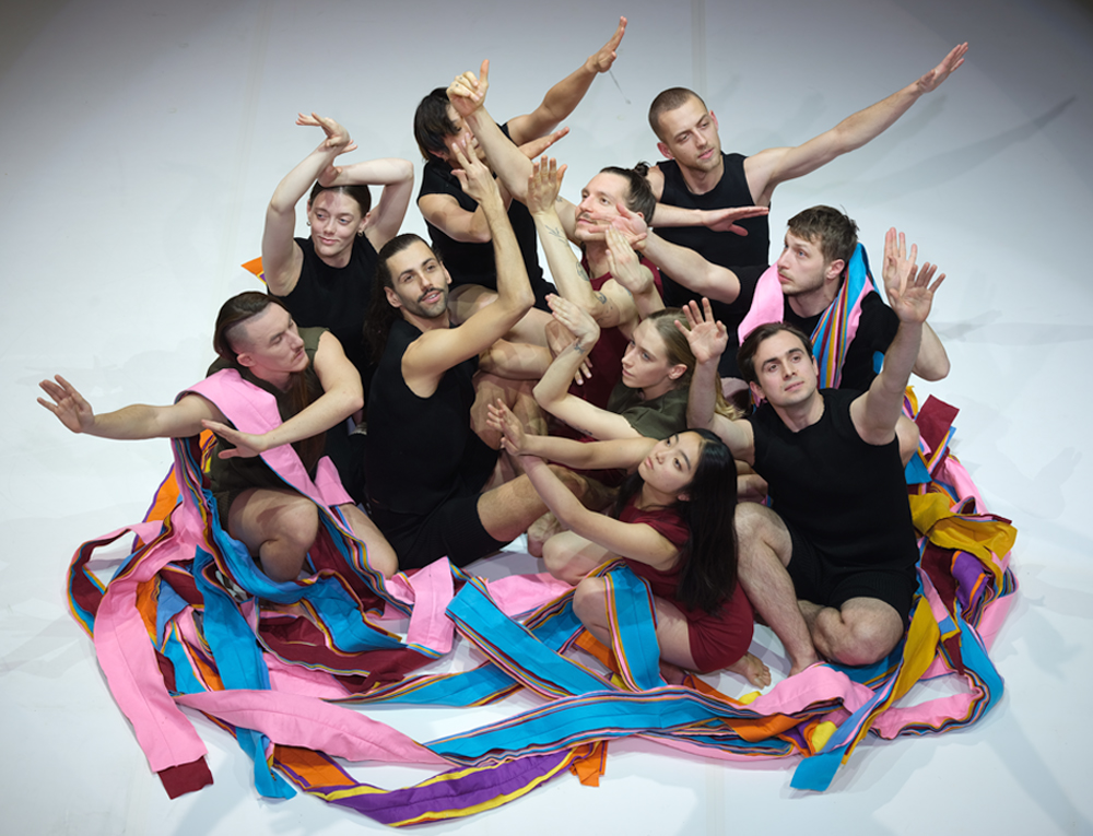 Zdjęcie grupy tancerzy siedzących na podłodze, z uniesionymi rękoma; niektórych z nich oplatają kolorowe szarfy.