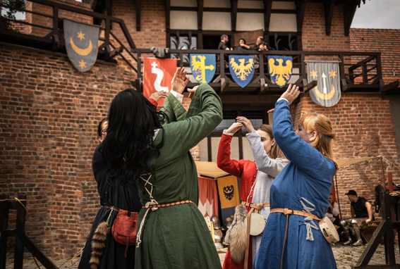 Grupa osób w średniowiecznych strojach tańczy na tle zamku i herbów.