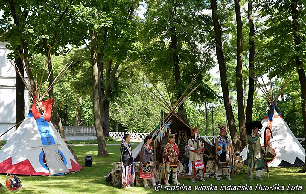 Indianie w charakterystycznych dla ich kultury strojach oraz namioty tipi. W tle trawa, drzewa i biały płot.