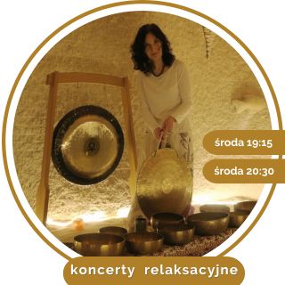 Kobieta w średnim wieku, w białym stroju, z czarnymi kręconymi włosami. Stoi przy rozłozonych złotych misach, trzyma duży gong w ręce. W tle wisi drugi gong.