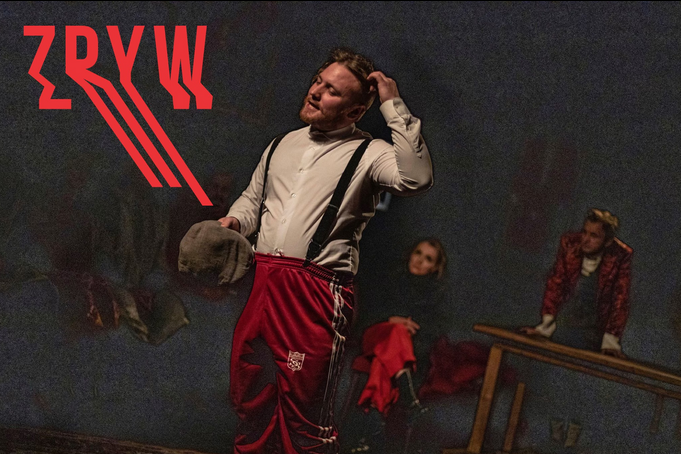 Zdjęcie przedstawia scenę teatralną w ciemnej tonacji. Na pierwszym planie stoi mężczyzna w białej koszuli z szelkami i czerwonych spodniach dresowych, trzymający w dłoni kaszkiet i drapiący się po głowie. W tle, w półmroku, widoczne są dwie inne postacie oraz fragment drewnianego stołu. W górnym lewym roku tytuł spektaklu zapisany czerwoną, niejako rozmytą czcionką.