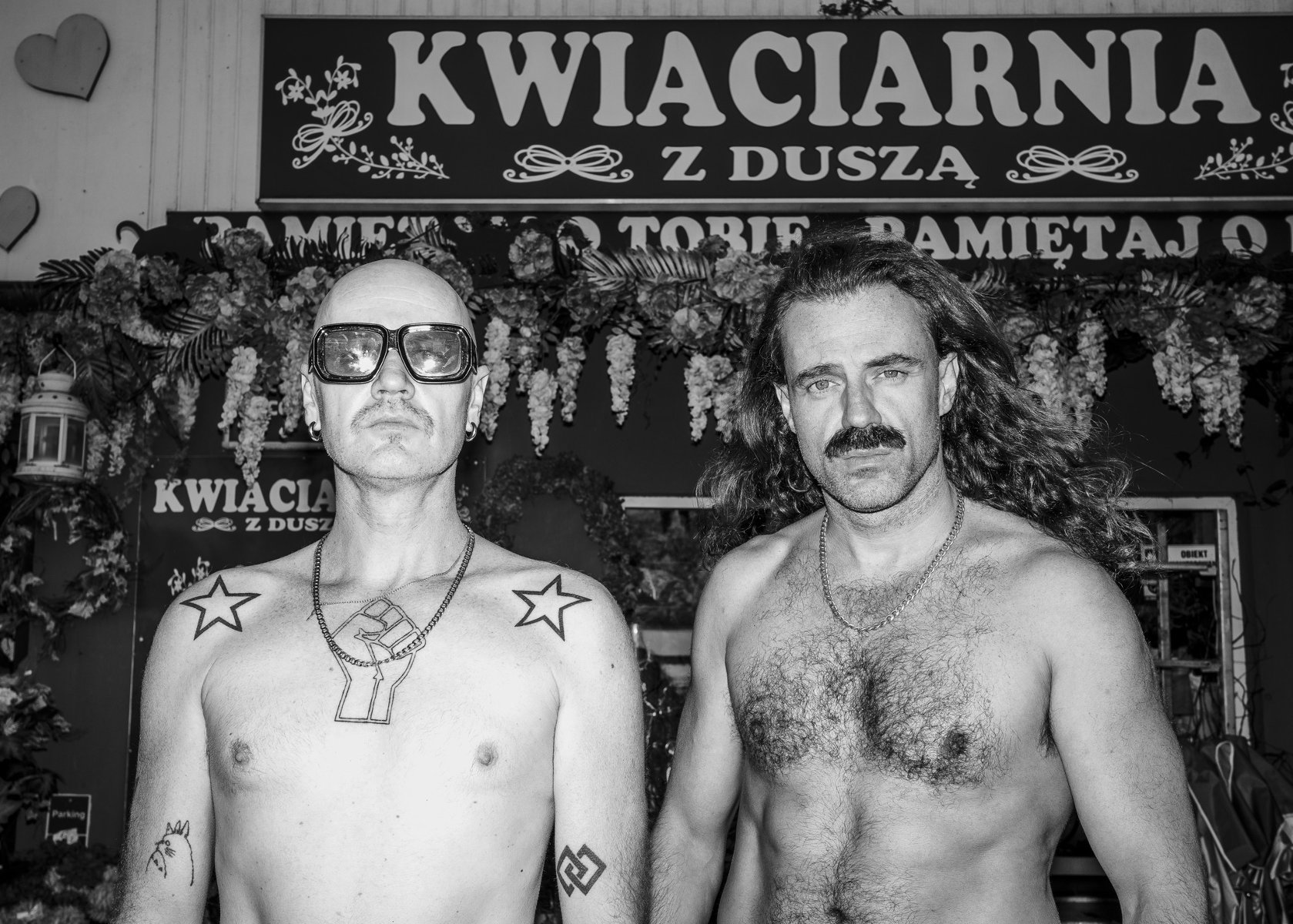 Czarno-białe zdjęcie przedstawia dwóch mężczyzn stojących przed kwiaciarnią z szyldem "Kwiaciarnia z duszą". Mężczyzna po lewej ma ogoloną głowę, ciemne okulary, tatuaże na klatce piersiowej i ramieniu. Mężczyzna po prawej ma długie włosy i wąsy. Obaj są bez koszulek.