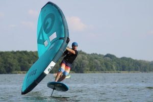 Zdjęcie osoby uprawiającej wingfoiling na jeziorze. Sportowiec, ubrany w kask i kolorowe spodenki, unosi się nad powierzchnią wody na desce. W dłoniach trzyma duży, turkusowy żagiel z ciemnymi detalami, który jest wygięty pod wpływem wiatru. W tle widać gęstą linię zielonych drzew na brzegu oraz jasne, pogodne niebo.
