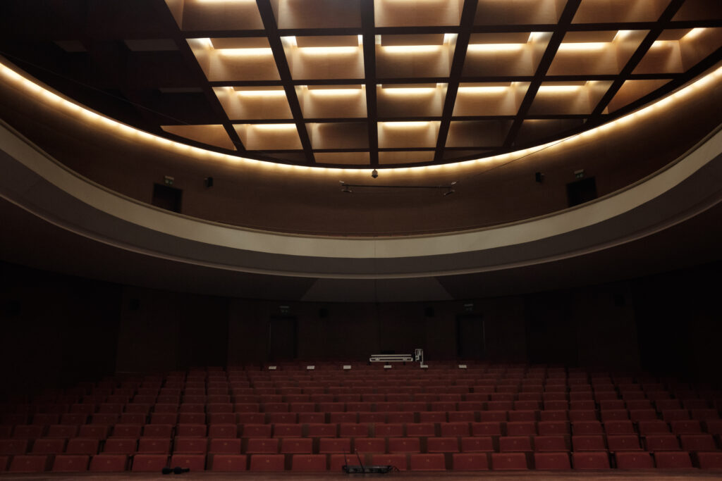 Obrazek przedstawia ciemną aule z czerwonymi krzesłami