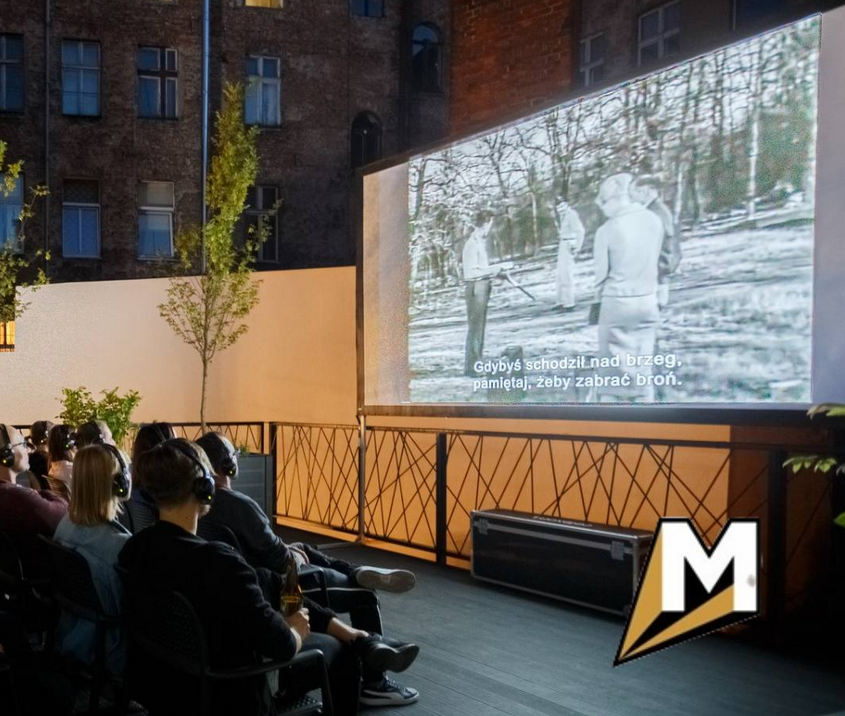 Zdjęcie, osoby w słuchawkach na uszach siedzą przed dużym ekranem na dachu kina.