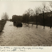 Powódź w mieście, ludzie płyną łodzią środkiem ulicy. Po prawej drzewa, po lewej płot.