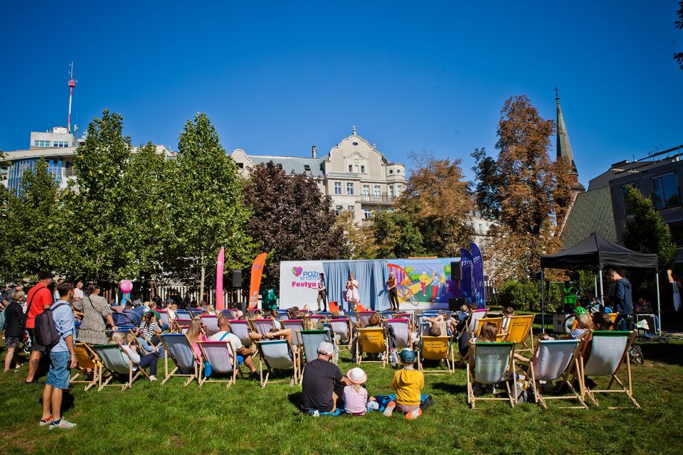Ludzie siedzą na zielonej trawie w parku. Patrzą na scenę, na której występują ludzie. Scena jest ozdobiona logo, kolorowymi rysunkami.