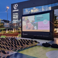 Przestrzeń przed budynkiem centrum handlowego, wieczór. Przed budynkiem ekran z wyświetloną reklamą wydarzenia "Kino letnie". Przed ekranem w rzędach ustawione leżaki, na kilku z nich siedzący ludzie.