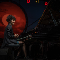 Przy fortepianie roześmiana pianistka jazzowa pochodzenia afrykańskiego, z burzą lokowatych włosów na głowie, siedząca bokkiem do widza.
