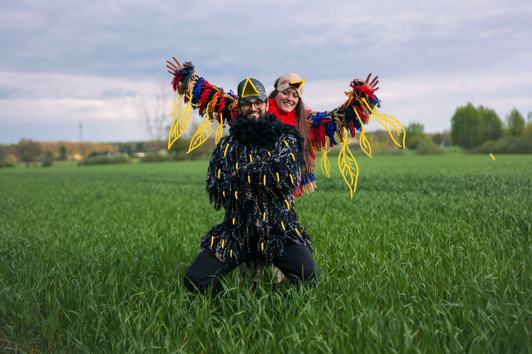 Kobieta i mężczyzna przebrani za kolorowe ptaki pozują do zdęcia. Stoją na zielonej polanie, za nimi widok na las.