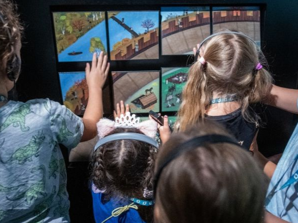 Grupa dzieci w słuchawkach dotyka kolorowej, interaktywnej wystawy w muzeum.