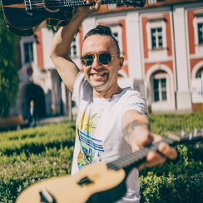 Rozmówca w białej koszulce na dziedzińcu Urzędu Miasta Poznania, trzyma w rękach dwa instrumenty ukulele, przypominające małe gitary