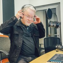 Ryszard Ćwirlej jest ubrany w czarną koszulkę i rozpinaną koszulę, zakłada sobie teraz na uszy słuchawski radiowe.