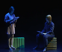 Zdjęce dwóch osób na scenie czytających tekst, jedna osoba stoi, druga osoba siedzi na skrzynce.