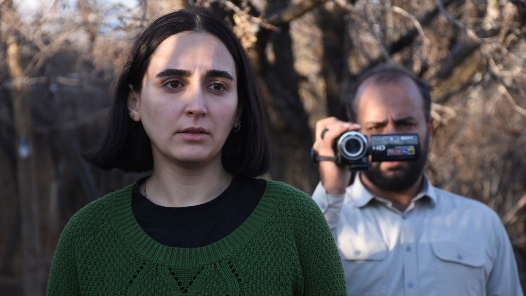 Młoda kobieta z ciemnymi włosami do ramion. Ubrana w czarną koszulkę i zielony sweter. Patrzy przejęta w dal. Za nią starszy mężczyzna z ciemnymi włosami i zarostem. Ubrany w jasną koszulę. W ręce trzyma kamerę cyfrową.