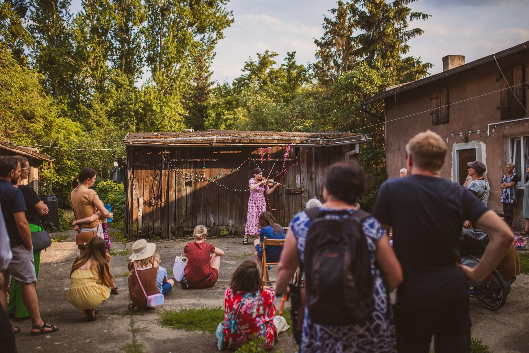 Tłum słucha kobiety grającej na skrzypcach na kameralnym podwórku domu.