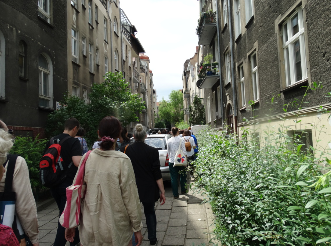 Uliczka między kamienicami, a na niej spacerująca grupa osób.