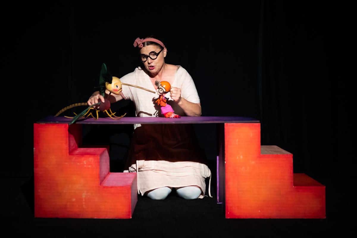 Zdjęcie ze spektaklu - aktorka klęcząc za elementem scenografii trzyma w rękach dwie lalki.