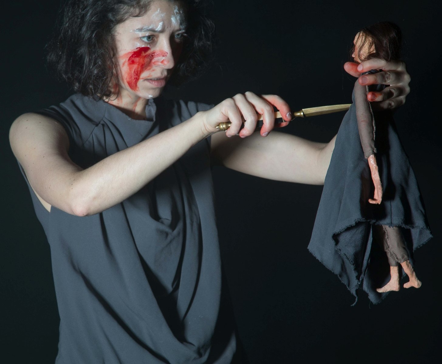 Zdjęcie ze spektaklu - kobieta z zakrwawioną twarzą w jednej ręce trzyma lalkę, a w drugiej sztylet, który przykłada do lalki.