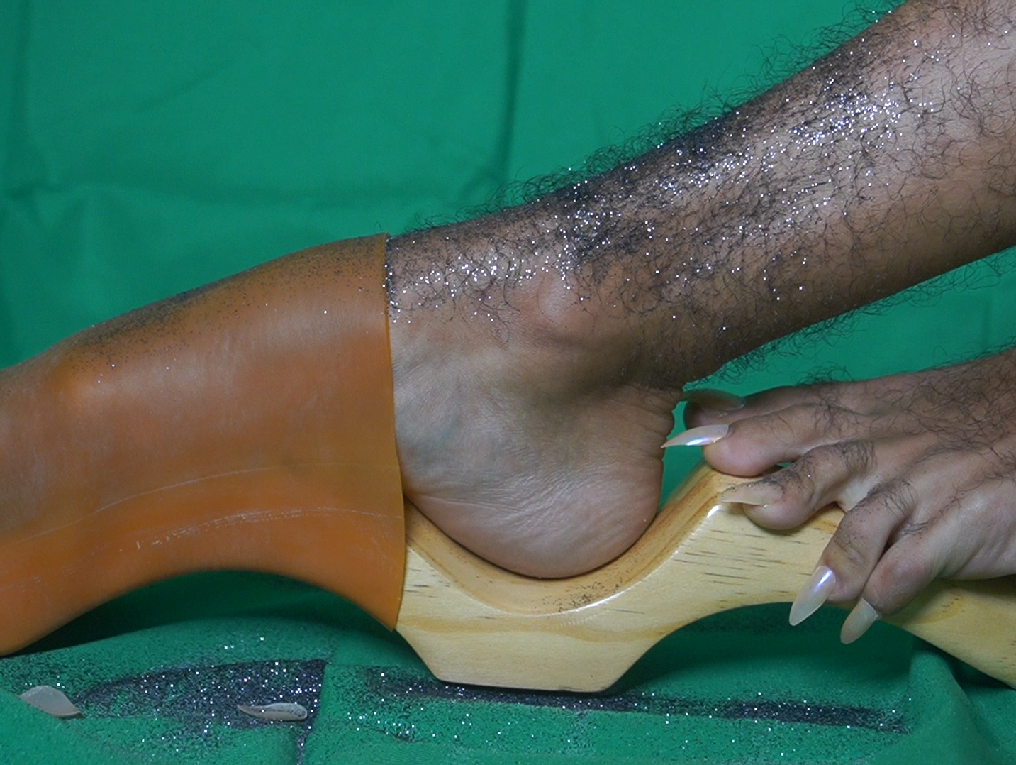 Zdjęcie owłosionej nogi pokrytej brokatem w drewnianym chodaku. Obok fragment drugiej stopy z długimi paznokciami.