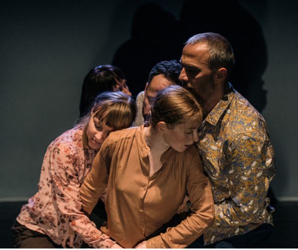 Zdjęcie przedstawia grupę aktorów w trakcie trwania spektaklu. Po lewej stronie znajdują się trzy kobiety, po prawej dwóch mężczyzn. W tle widoczne są dwie nieoświetlone sylwetki, sprawiające wrażenie kryjących się w cieniu.