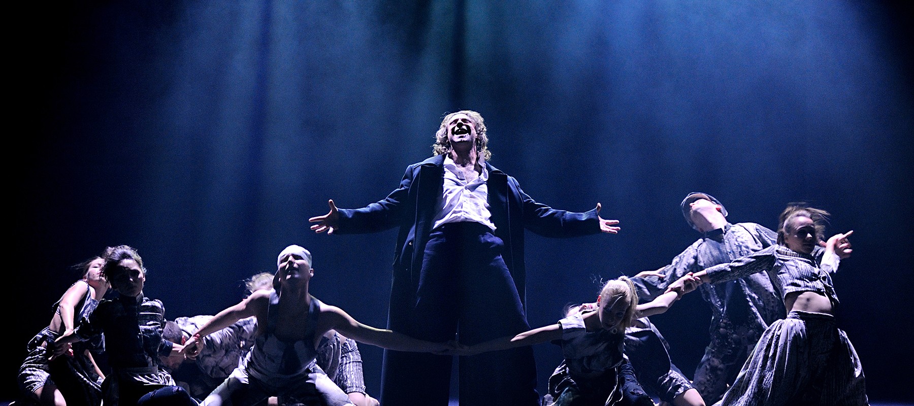 Zdjęcie ze spektaklu - główny bohater śpiewa na scenie w otoczeniu tancerzy.