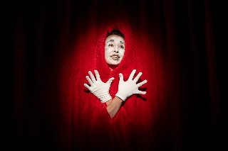 Postać w czerwonym stroju i wymalowaną na biało twarzą stoi na tle kurtyny teatralnej.