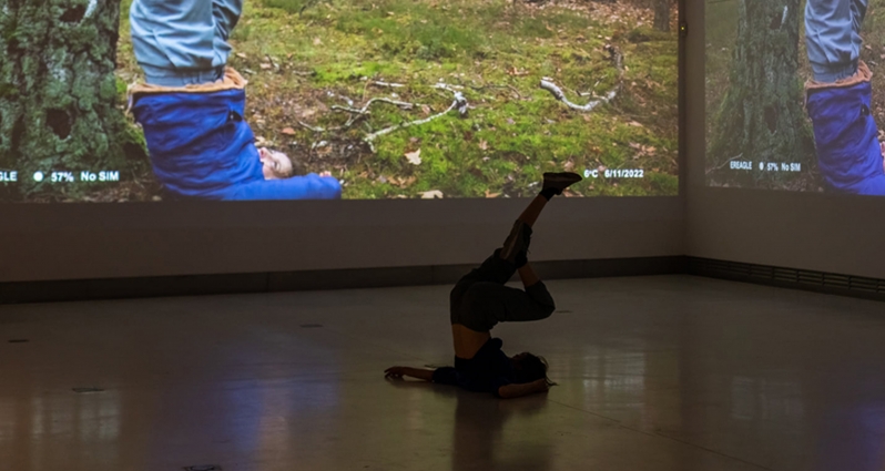 Zdjęcie ze spektaklu - tancerka leżąca na podłodze z uniesionymi pionowo nogami. W tle fragment zdjęcia wyświetlonego na ścianie, przedstawiającego osobę w podobnej pozie na trawie przy drzewie.