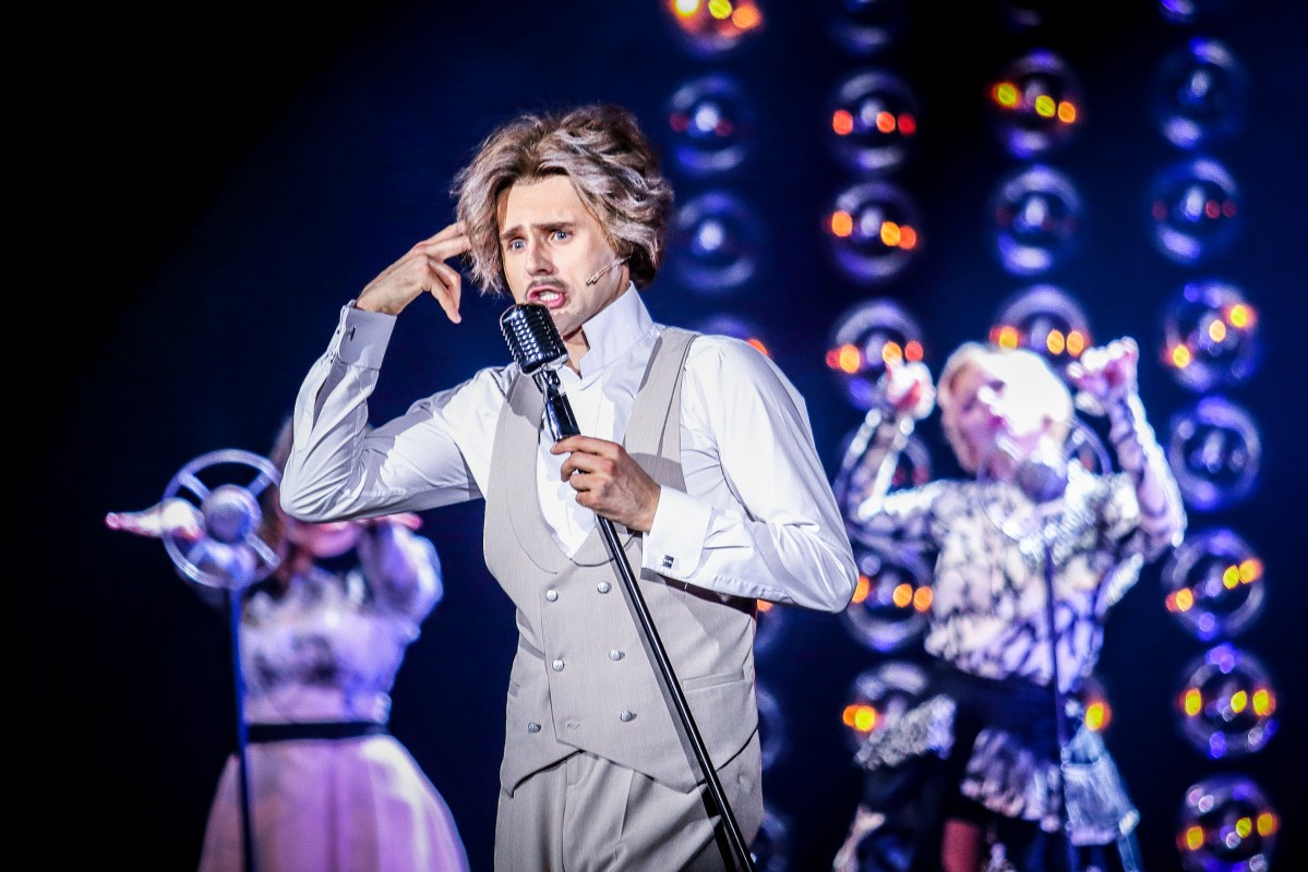 Zdjęcie ze spektaklu - mężczyzna w bialej koszuli i szarych kamizelce i spodniach śpiewający do mikrofonu. W tle dwie aktorki przy mikrofonach.