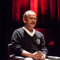 Starszy łysiejący mężczyzna z haryzmą oczach i wąsem siedzi z mikrofonem na tle kurtyny teatralnej.