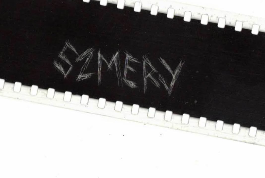 Grafika przypomina otwarty zamek błyskawiczny. W otwartej przestrzeni widać czarne tło z wydrapanym napisem "szmery".