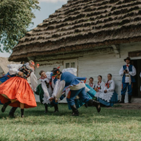 Tancerze w polskich strojach ludowych tańczą przed tradycyjną, krytą strzechą chatą chłopską.