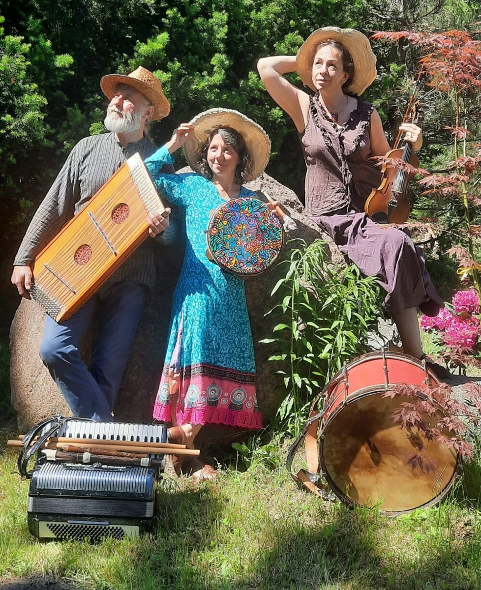 Mężczyzna i kobiety w średnim wieku ubrani w kolorowe stroje. Na głowach mają słomkowe kapelusze a w rękach trzymają różne instrumenty muzyczne. Przed nimi leżą inne instrmenty. Za nimi drzewa i kolorowe kwiaty.