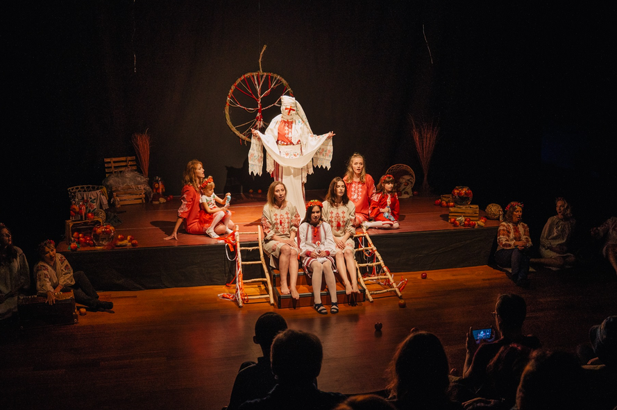 Grupa osób w słowiańskich strojach z wzorami ludowymi siedzi na drewnianej scenie teatralnej.