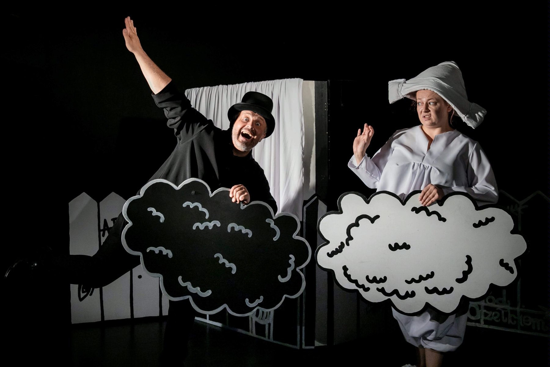 Zdjęcie, na czarnym tle, stoją aktorzy w czarno - białych strojach i trzymają w rękach czarne i białe chmurki.