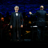 Tenor śpiewa do mikrofonu, za nim dyrygent i orkiestra.