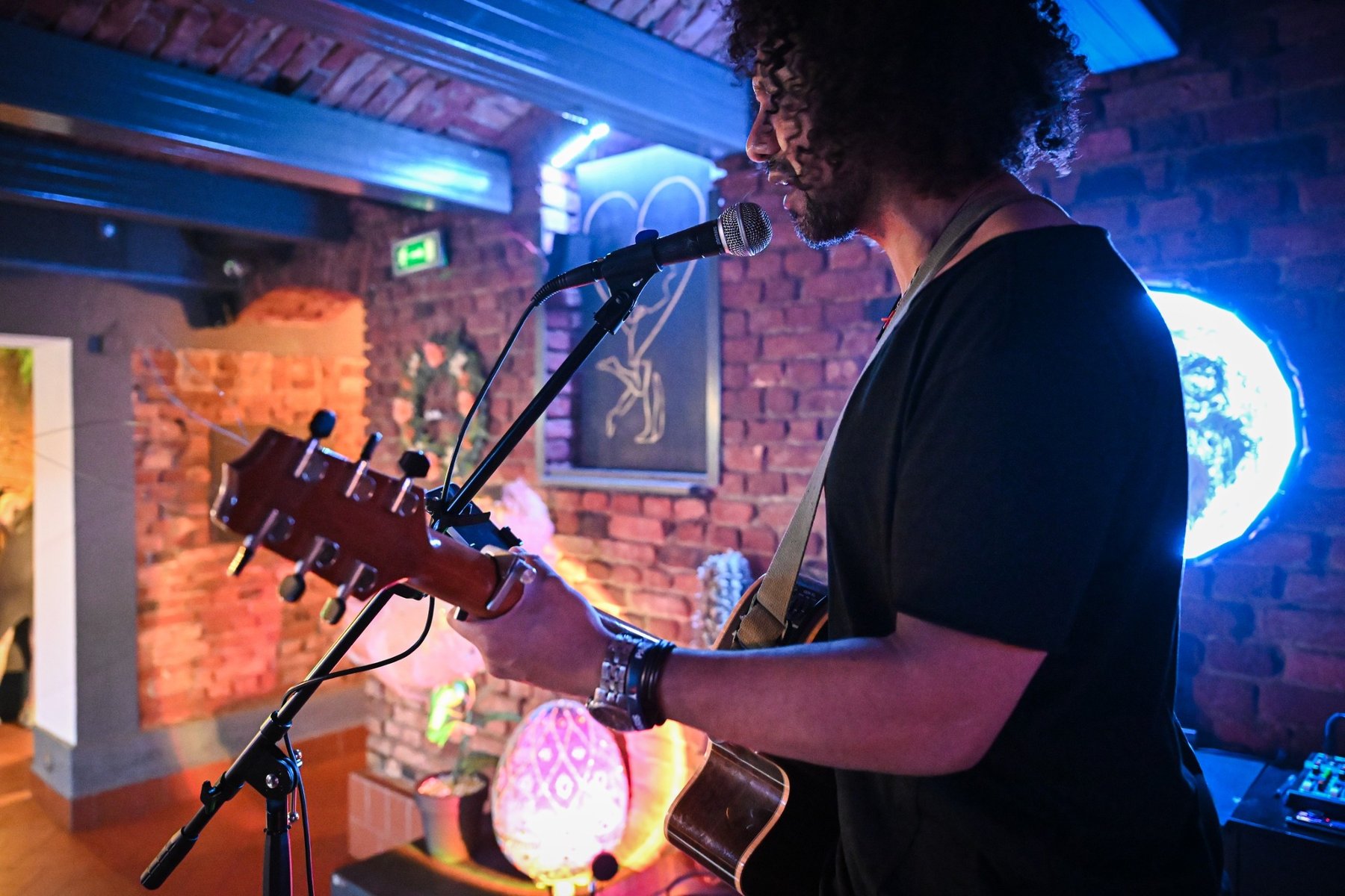Mężczyzna z kręconymi włosami. Trzyma w rękach gitarę. Śpiewa do mikrofonu. W tle ceglana ściana z ozdobami.