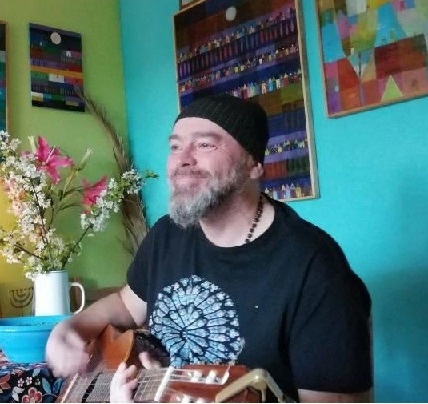 Tomasz Budzyński w czarnej czapce i ciemnej koszulce z gitarą w mieszkaniu. Po lewej zielona ściana, po prawej turkusowa. Na ścianie obrazy.