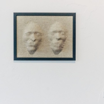 Trójwymiarowy obraz wiszący na ścianie. Przedstawia dwie ludzkie twarze, jakby wypchnięte przez beżową, przypominającą len tkaninę.