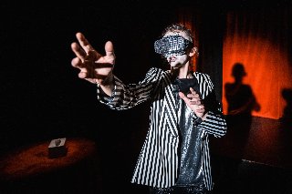 Na zdjęciu stoi kobieta w garniturze w paski. Jej ramię wyciąga się w stronę publiczności. Za nią znajduje się czerwona scena za kulisami.