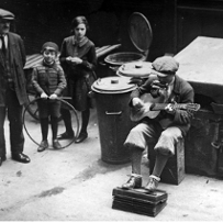 W centrum czarno-białej fotografii jest muzyk z gitarą. Słucha go starszy meżczyna, młoda kobiea i dziecko. Muzyk siedzi obok metalowych kubłów na śmieci.