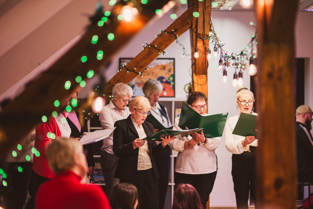 Grupa śpiewającycj osób w udekorowanym świątecznie pomieszczeniu.