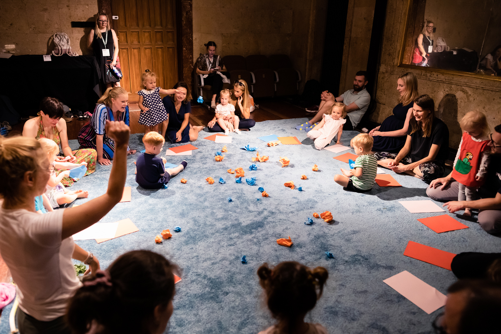 Zdjęcie sali, w której na podlodze pokrytej jasno niebieskim materiałem siedzą dzieci, przy nich rodzice. Na środku rozrzucone niebieskie i pomarańczowe zgniecione kawałki papieru.