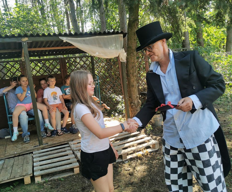 zdjęcie dziewczynki podającej rękę mężczyźnie we fraku i kapeluszu, w tle dzieci siedzące w altanie