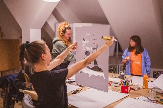 Trzy kobiety na warsztatach plastycznych. Jedna ogląda portret, dwie stoją przy stole z rozłożonymi materiałami plastycznymi (pędzle, kartki, farby).