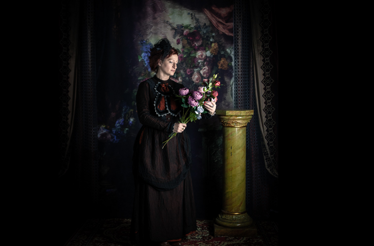 Zdjęcie, na ciemnym tle stoi kobieta w długiej sukni, trzymająca bujkiet kwiatów.