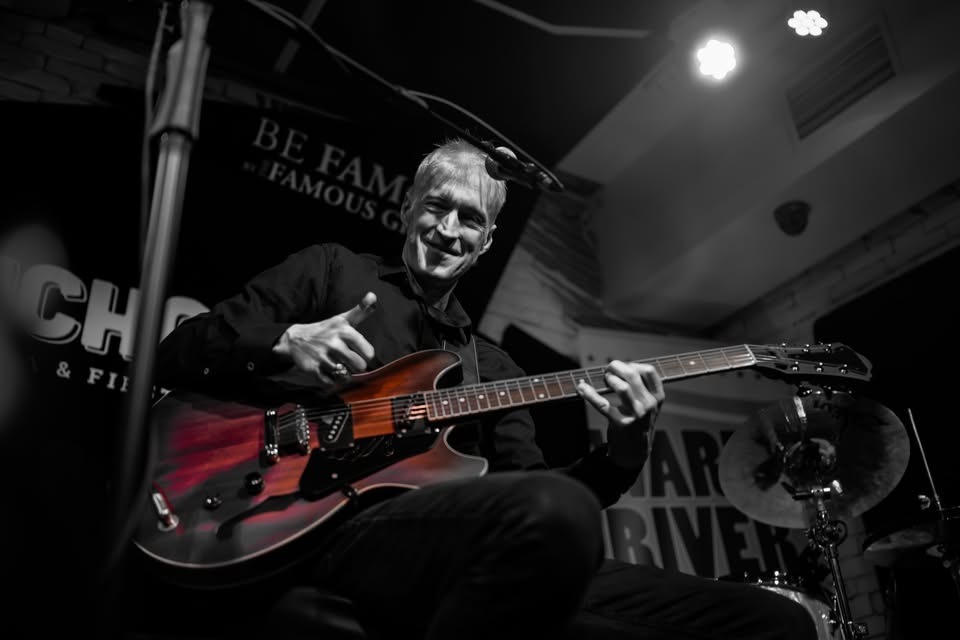 Mężczyzna na scenie pokazuje gest kciuka w górę grając na gitarze.