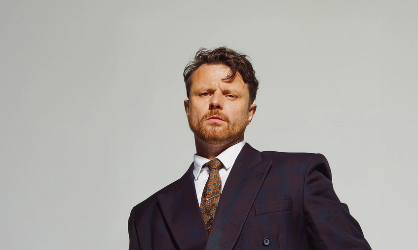 Artysta w garniturze i krawacie na tle szarej ściany.